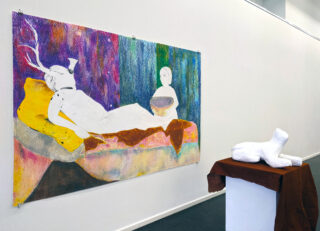Farbenfrohes Gemälde zweier abstrakter Figuren, von denen eine auf einem Bett liegt und die andere in der Nähe sitzt. Sie werden neben einer weißen abstrakten Skulptur auf einem braun bedeckten Sockel ausgestellt.