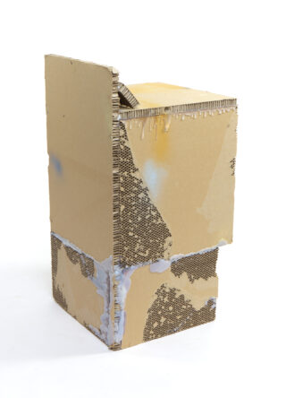 Eine rechteckige Skulptur aus Wellpappe mit rauen Kanten, teilweise bedeckt mit Leimtropfen und Farbklecksen, steht aufrecht vor einem weißen Hintergrund.
