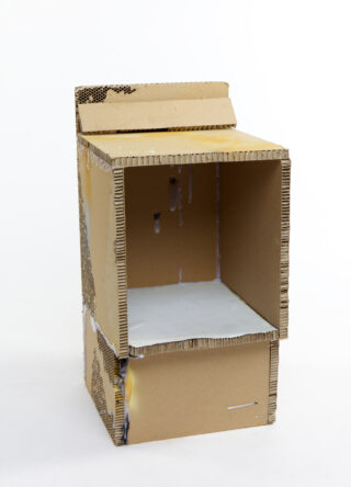 Eine rechteckige Kartonstruktur mit offener Vorder- und Oberklappe, mit einigen Tropfen weißer Farbe und sichtbarer Riffelung an den Kanten.