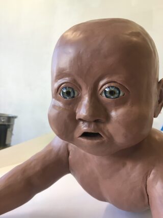 Eine realistische Ton-Skulptur eines Babys mit blauen Glasaugen, von den Schultern aufwärts vor einem schlichten Hintergrund dargestellt.