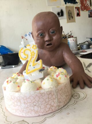 Eine lebensechte Babypuppenskulptur steht hinter einem Kuchen, der mit einer Kerze mit der Zahl 2 und weißem Zuckerguss verziert ist, auf einem Tisch im Atelier eines Künstlers.