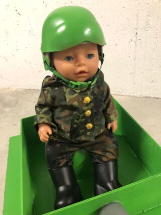 Eine Puppe in Tarnkleidung und mit grünem Helm sitzt in einer grünen Schachtel vor einem schlichten Hintergrund.