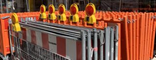 Auf einer Baustelle sind Reihen orangefarbener und weißer Bauabsperrungen mit daran befestigten gelben Warnlichtern übereinander gestapelt.