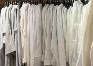 Eine Reihe weißer und hellgrauer Hemden hängen auf schwarzen Kleiderbügeln in einem Schrank.