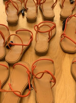 Mehrere Paar hellbraune Sandalen mit orangefarbenen Riemen sind auf einem Holzboden angeordnet und zeigen alle in unterschiedliche Richtungen.