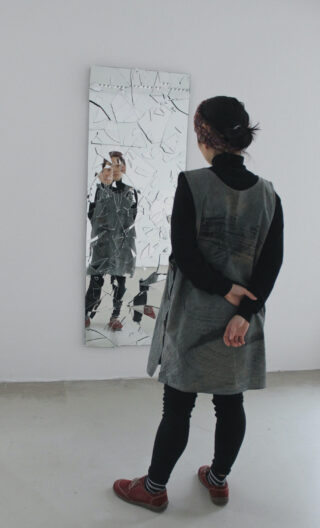 Eine Person steht vor einem großen, zerbrochenen Spiegel. Ihr zersplittertes Spiegelbild ist im zerbrochenen Glas sichtbar.