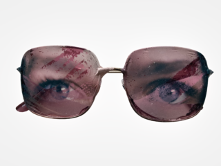 Eine rosa getönte Sonnenbrille mit Wassertropfen auf den Gläsern, die die Reflexion menschlicher Augen zeigen, die hindurchblicken.