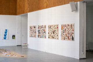 Eine Galerie für zeitgenössische Kunst mit vier großen Collagen an einer weißen Wand, einem einzelnen blauen Kunstwerk an der Seite, verstreuten Gegenständen auf dem Boden und einem kleinen Hocker in der Nähe.