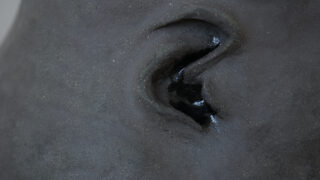 Nahaufnahme einer strukturierten dunkelgrauen Skulptur, die ein verzerrtes menschliches Ohr mit einer glänzenden schwarzen Substanz im Gehörgang zeigt.