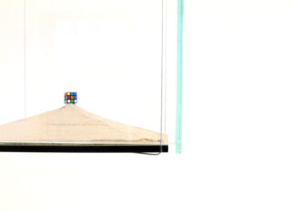 Ein Zauberwürfel liegt auf einem kleinen Sandhügel in einer Glasvitrine vor einem schlichten weißen Hintergrund.