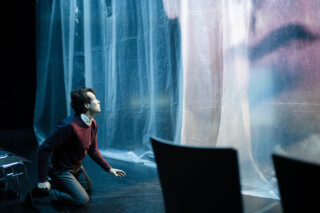 Eine Person kniet auf dem Boden und blickt auf eine durchsichtige Plastikfolie, auf die ein großes, verschwommenes Gesicht projiziert wird; im Vordergrund sind Stühle zu sehen.