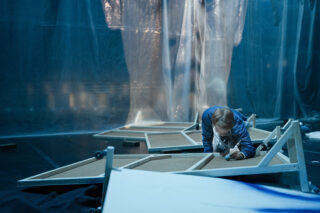 Eine Person im blauen Overall kauert auf dem Boden und streckt die Hand nach einer eingestürzten Holzkonstruktion aus, im Hintergrund hängen durchsichtige Plastikfolien.