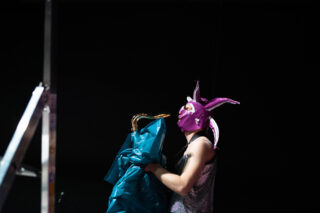 Eine Person in einem glänzenden Outfit und einer lila Hasenmaske hält vor einem dunklen Hintergrund ein glänzendes blaues Material neben einer Leiter.