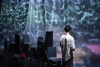 Eine Person steht vor einer strukturierten, beleuchteten Wand in der Nähe mehrerer gestapelter Stühle; auf einem der Stühle liegt ein roter Gegenstand.