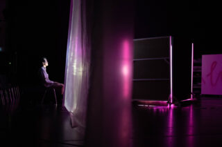 Eine Person sitzt auf einem Stuhl vor einem Bildschirm oder einer beleuchteten Tafel mit rosa und violetter Beleuchtung, die teilweise durch einen durchscheinenden Vorhang in einem dunklen Raum verdeckt wird.