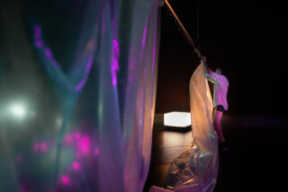 Eine Person in einem weißen Hemd richtet ein großes transparentes Laken auf einer schwach beleuchteten Bühne mit farbigen Lichtern und einem leuchtenden Würfel im Hintergrund.