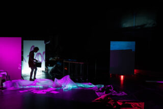 Eine Person steht in einem schwach beleuchteten Raum mit bunter Beleuchtung, umgeben von verstreuten Papieren, Stoffen und durchscheinendem Material auf dem Boden.