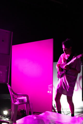 Eine Person steht in einem magentafarben beleuchteten Raum und spielt E-Gitarre. Um sie herum sind ein Plastikstuhl, Paneele und verschiedene Geräte zu sehen.
