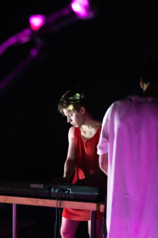 Eine Person in einem roten Outfit spielt auf der Bühne ein Yamaha-Keyboard. Eine weitere Person in einem weißen Hemd steht daneben. Lila Bühnenbeleuchtung ist zu sehen.