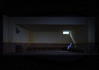 Eine weiß gekleidete Person sitzt allein auf einer Bank in einem schwach beleuchteten, größtenteils leeren Raum mit Holzvertäfelung und einem kleinen Fenster, das Licht hereinlässt.