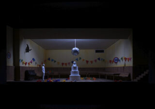 Ein schwach beleuchteter Raum mit einer großen mehrstöckigen Torte in der Mitte, zwei Schaufensterpuppen, bunten Wimpelketten, Luftballons und einer Discokugel, die von der Decke hängt.