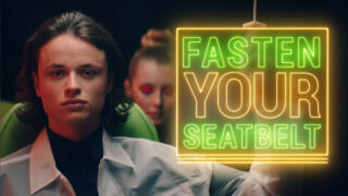 Eine Person sitzt in einem Flugzeug, hinter ihr ein weiterer Passagier, neben einem hellen Neonschild mit der Aufschrift „ANSCHNELL ANSCHNELLT“.