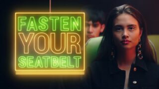 Auf einem Neonschild steht „ZAHNSCHNALLE“ neben einer Frau mit Gesichtspiercings und Ohrringen, die auf einem Sitz sitzt, im Hintergrund ist eine Person zu sehen.