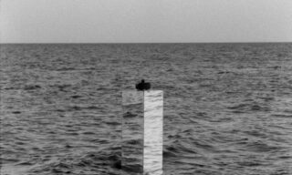 Eine Person sitzt auf einer hohen, rechteckigen, marmorartigen Struktur, umgeben von offenem Wasser unter einem bewölkten Himmel.