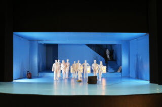 Eine Gruppe weißer Schaufensterpuppen steht auf einem blauen Bühnenbild mit Treppen im Hintergrund und mehreren Requisiten, die um sie herum platziert sind.