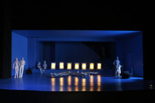 Ein Bühnenmodell zeigt sechs weiße menschliche Figuren, aufgerollte Kabel und eine Reihe beleuchteter rechteckiger Lichter unter blauer Beleuchtung.