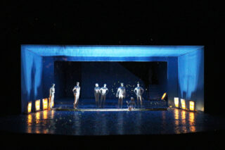 Eine Bühnenszene mit sieben silbernen menschlichen Figuren, die unter einer blauen rechteckigen Struktur stehen, die von blauen und gelben Lichtern auf einem reflektierenden Boden beleuchtet wird.