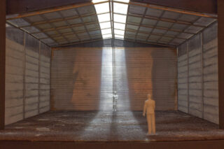 Zwei kleine weiße Figuren stehen in einem großen, leeren Lagerhaus. Durch ein Oberlicht fällt Licht auf eine Figur in der Mitte.
