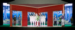 Ein Modell eines roten Backsteingebäudes mit Papierausschnitten von Menschen, darunter eine Gruppe von Tänzern in Weiß, die vor großen Fenstern und einer Waldlandschaft im Hintergrund angeordnet sind.