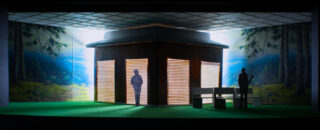 Ein sechseckiges Gebäude mit beleuchteten Fenstern steht auf Kunstrasen, mit zwei menschlichen Silhouetten und Bänken in der Nähe, vor einer Kulisse mit Waldmotiven.