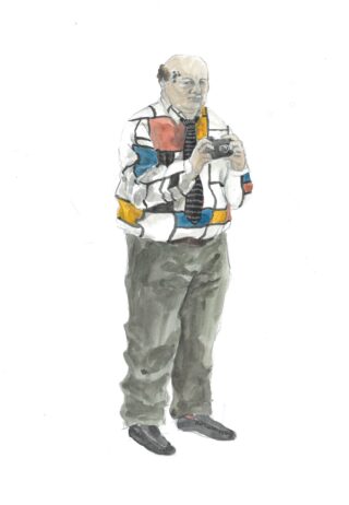 Ein Mann mit Glatze und einem Hemd mit geometrischem Muster hält eine Kamera und blickt nach vorne.