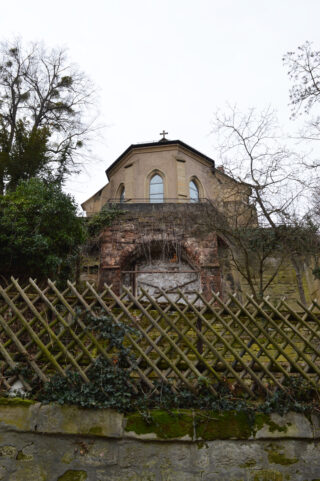 Eine beige Kirche mit Bogenfenstern und einem Kreuz darauf wird teilweise von einem hölzernen Gitterzaun, überwucherten Pflanzen und kahlen Bäumen verdeckt.
