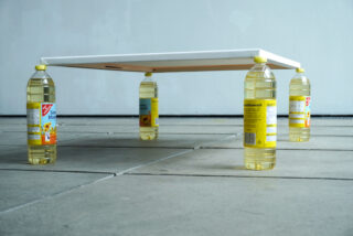 Ein rechteckiges Holzbrett wird durch vier mit gelber Flüssigkeit gefüllte Plastikflaschen erhöht, eine an jeder Ecke, und dient als improvisierte Tischbeine auf einem gefliesten Boden.