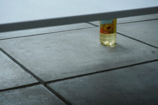 Eine Flasche Sonnenblumenöl dient dazu, die Kante eines weißen Tisches auf einem gefliesten Boden abzustützen.