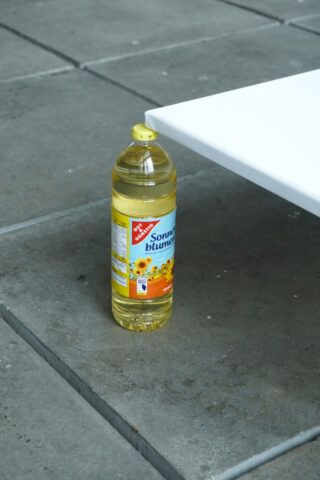 Eine Plastikflasche Sonnenblumenöl mit gelbem Deckel steht auf einem gefliesten Boden neben einer weißen rechteckigen Fläche.