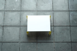 Von oben betrachtet steht ein kleiner weißer rechteckiger Tisch mit gelben Beinen auf einem grau gefliesten Boden.