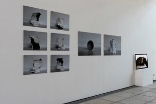 Eine Reihe Schwarzweißfotos von gestapelten Papieren und Objekten wird an einer weißen Galeriewand angezeigt, während rechts auf einem Sockel ein gerahmtes Foto steht.