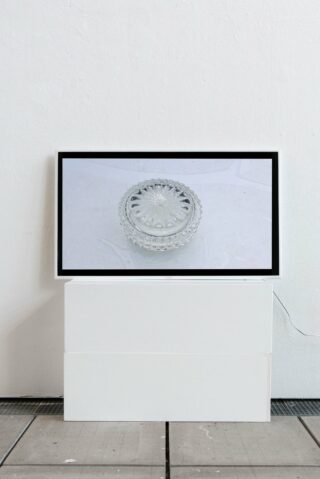 Ein Flachbildschirm, auf dem das Bild einer durchsichtigen Glasschale zu sehen ist, steht auf zwei weißen rechteckigen Blöcken vor einer schlichten weißen Wand.