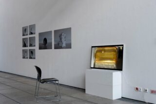 Ein schwarzer Stuhl steht vor einer Wand, auf der sechs Graustufenfotos und ein Monitor mit einem gelben Objekt zu sehen sind – alles in einem minimalistischen Galerieraum.