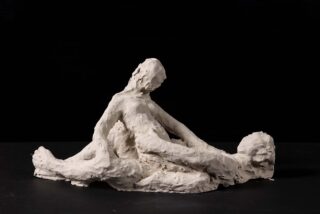 Eine Skulptur aus weißem Ton mit rauer Textur stellt vor einem schwarzen Hintergrund zwei abstrakte menschliche Figuren dar, von denen eine aufrecht sitzt und die andere auf ihrem Schoß liegt.