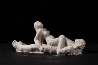 Eine Skulptur aus weißem Ton mit rauer Textur, die zwei abstrakte menschliche Figuren darstellt, die eng beieinander vor einem schwarzen Hintergrund liegen.