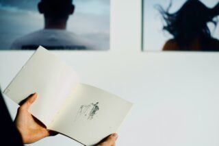 Eine Person hält vor einer Wand, an der zwei unscharfe Porträtfotos hängen, ein Skizzenbuch mit einer einfachen abstrakten Zeichnung aufgeschlagen.