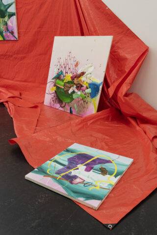 Zwei abstrakte Gemälde, eines an die Wand gelehnt und eines flach auf dem Boden liegend, werden auf einer leuchtend roten Plane in einer Galerieumgebung ausgestellt.