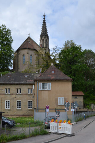 Hinter einem beigen Gebäude steht eine Kirche mit hohem Turm, im Vordergrund sind Bauabsperrungen und Verkehrsschilder zu sehen.