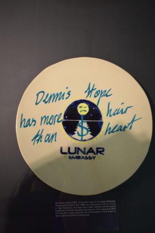 Ein Keramikteller mit dem Logo der Lunar Embassy und dem handgeschriebenen Satz „Dennis Hope hat mehr Haare als Herz“. Unter dem Teller ist ein kleines Beschreibungsetikett sichtbar.