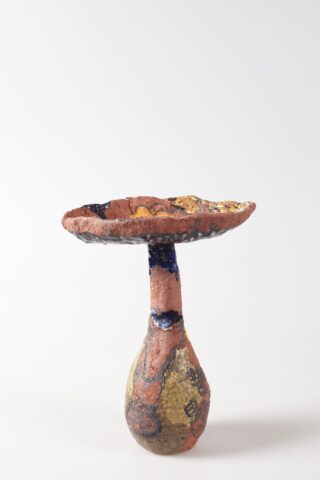 Ein skulpturales Keramikstück mit einer breiten, flachen Schale auf einem schmalen Sockel und strukturierten, mehrfarbigen Oberflächen in erdigen und dunklen Tönen vor einem schlichten weißen Hintergrund.
